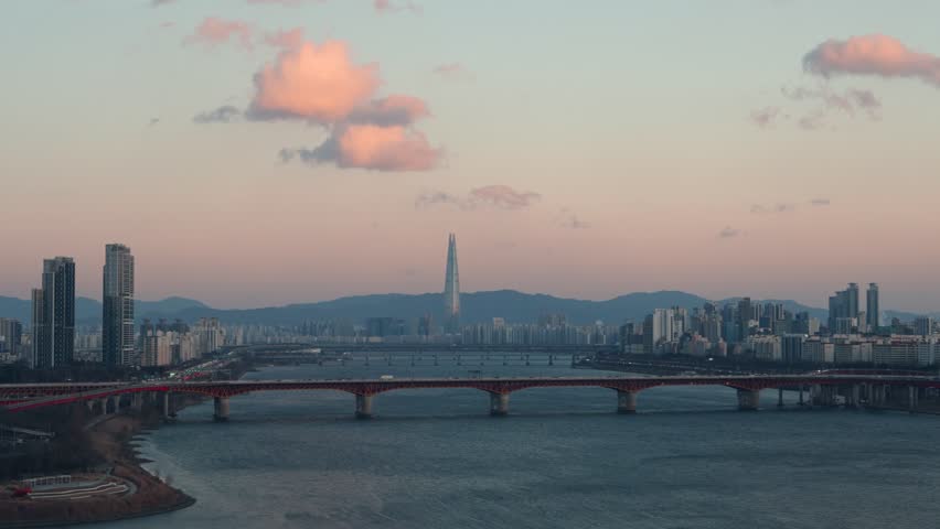 Aerial view captures Seoul skyline featuring Lotte World Tower and Seongsu Bridge over Han River at sunset with pink fluffy clouds and city traffic below on a winter evening