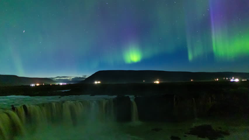 Northern lights at Godafoss Waterfall, Iceland. Dramatic glowing green and red columns in sky with car traffic lights and waterfall in foreground. Time lapse.