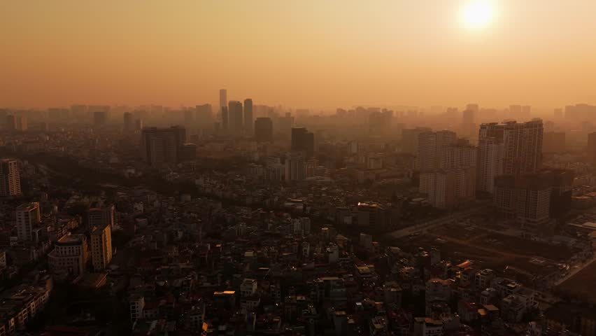 Drone ascends and pans over Hanoi skyline at sunset, showing dense buildings fading into haze in Vietnam