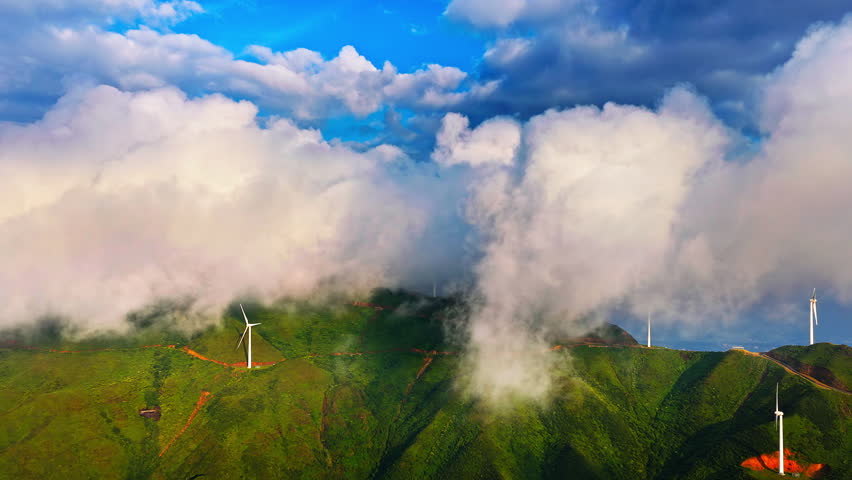 Aerial shot of modern wind turbines on green mountain ridge surrounded by thick white clouds under blue sky