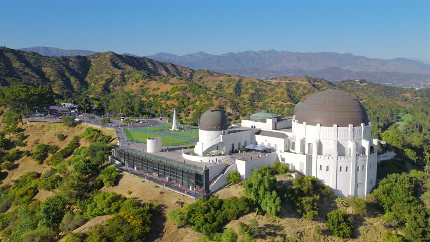 Aerial view of Griffith Observatory surrounded by green hills and mountains above Los Angeles California, iconic landmark overlooking the city skyline under clear blue sky