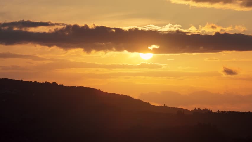 Sun setting behind heavy cloud layer over rolling hills, aerial silhouette landscape during golden hour, peaceful nature scene in Greece, romantic dusk sky with warm glow