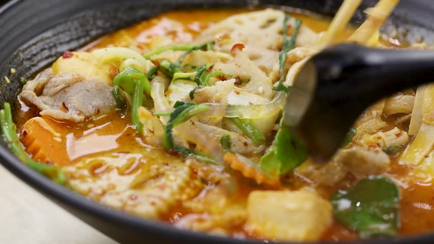 A close-up view of chopsticks picking up transparent noodles from a vibrant, spicy broth.