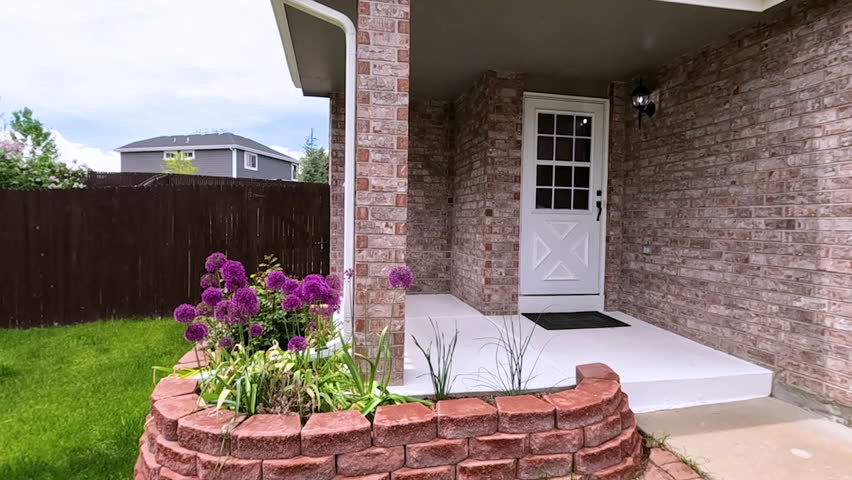 The front porch of a charming brick house featuring a white door and a well-maintained garden bed with blooming purple flowers. The neatly trimmed lawn and inviting entryway enhance the homes curb