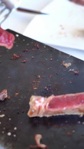 A slab of meat is on a black surface with a knife and fork nearby. The meat appears to be well-cooked and has been cut into pieces.
