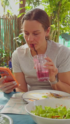 Vertical video. Young Woman Drinking Berry Smoothie Through Straw While Using Smartphone At Outdoor Cafe After Meal Showing Healthy Lifestyle And Modern Daily Routine.