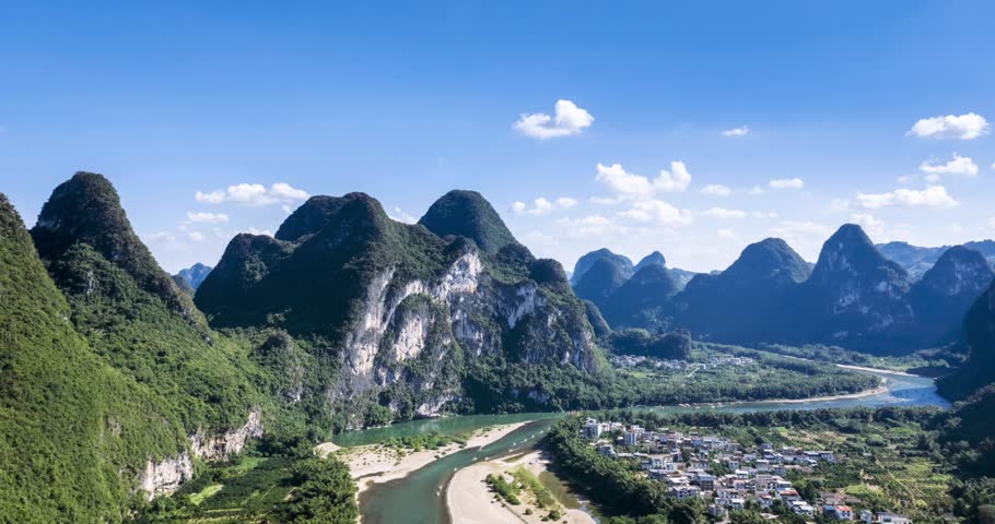 Aerial time-lapse of the Li River winding through dramatic karst peaks in Guilin, China. The iconic Nine-Horse Painting Mountain rises majestically under a clear blue sky, with lush greenery and a riverside village below.