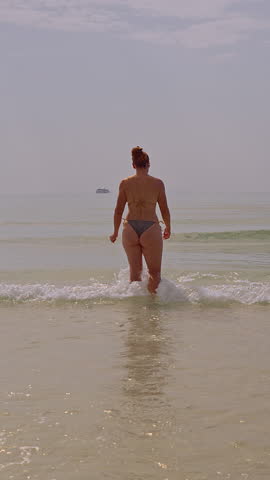 Vertical video. A Woman Stands in Shallow Sea Water Facing the Horizon as Soft Sunlight Reflects Across Gentle Waves Creating a Calm Peaceful Tropical Beach Atmosphere.