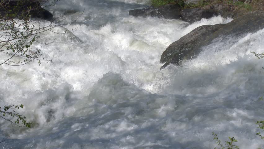 Super slow motion foamy white river water crashes through rocky mountain forest wild. Extreme detail reveals turbulent liquid energy and mist within a powerful natural scene.
