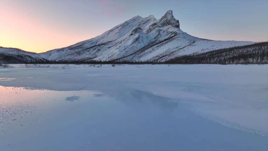 Aerial view of snow-covered mountains and frozen lake under a pastel sky, creating a serene and expansive winter landscape, Dalton Highway, Alaska, United States.