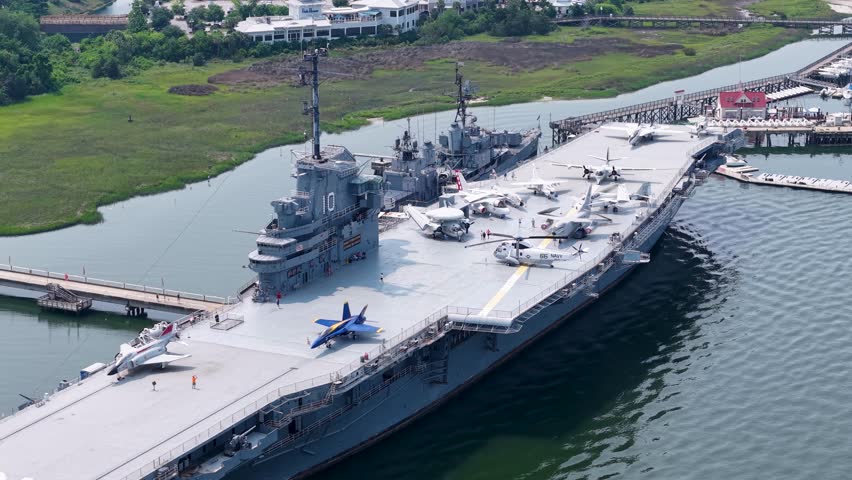 Drone footage captures an aerial side view of an aircraft carrier docked at a harbor with green water and surrounding facilities in Charleston, South Carolina, showing the large warship and its moorin