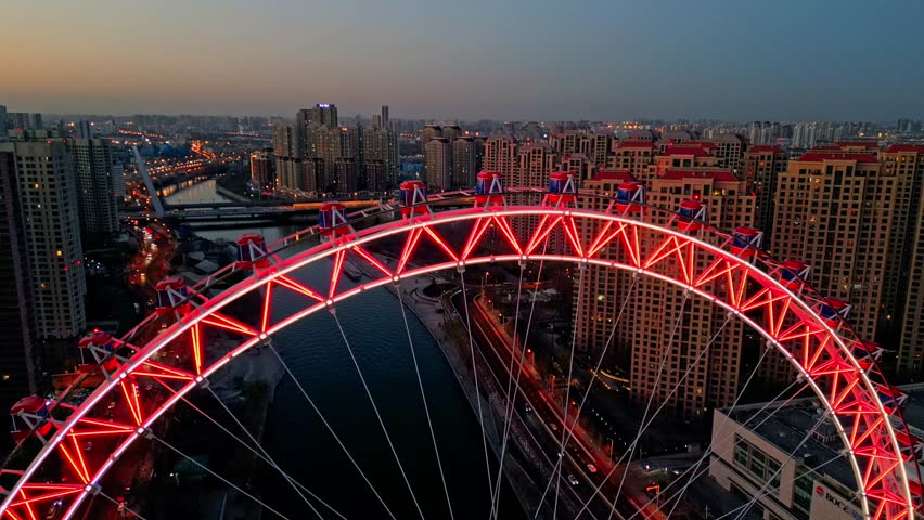 Aerial view of the illuminated Tianjin Eye ferris wheel against a backdrop of residential buildings and a winding river at dusk, Tianjin, Tianjin, China.
