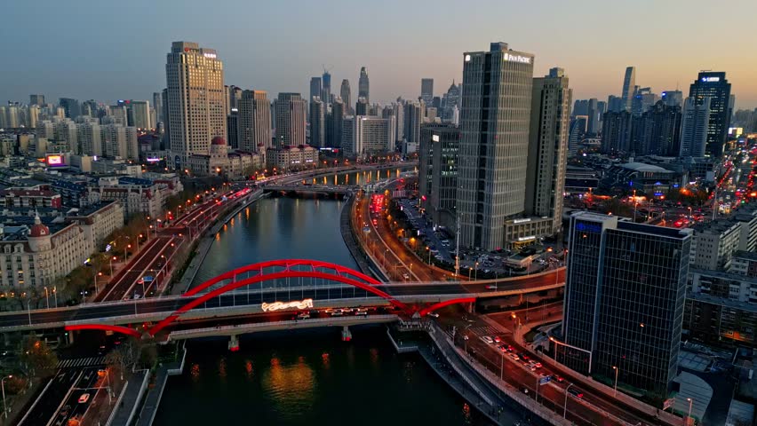 Aerial view of cityscape featuring a red bridge spanning a river, surrounded by buildings and illuminated roads, Tianjin, Tianjin, China.