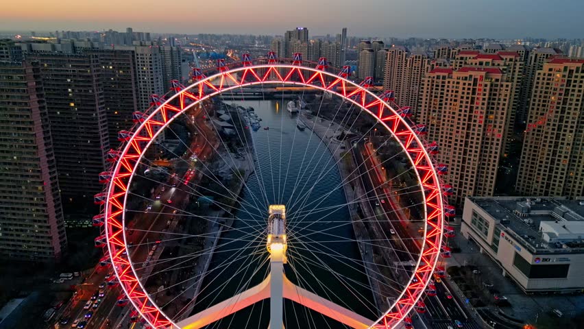 Aerial view of the brightly lit Tianjin Eye Ferris wheel towering over the city and river, its red lights contrasting against the buildings, Tianjin, Tianjin, China.