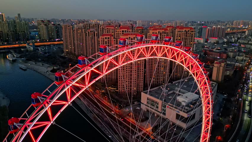Aerial view of the brightly lit Tianjin Eye Ferris wheel casting a warm red glow over the cityscape and river at dusk, Tianjin, Tianjin, China.