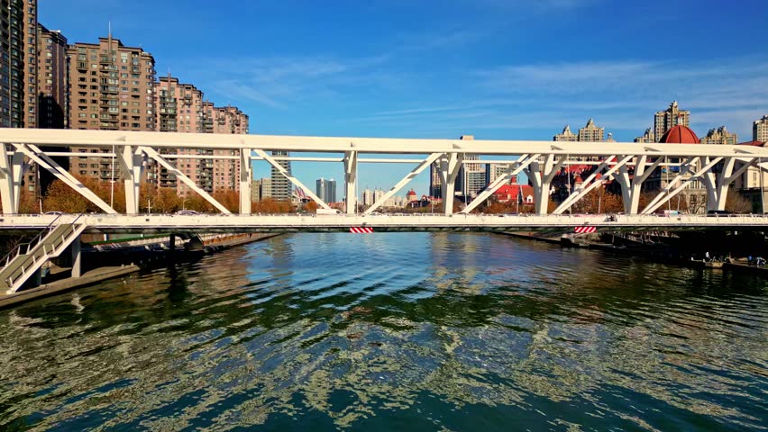 Aerial view of a white bridge over a river, reflecting the blue sky, surrounded by modern buildings and autumnal trees, Tianjin, Tianjin, China.