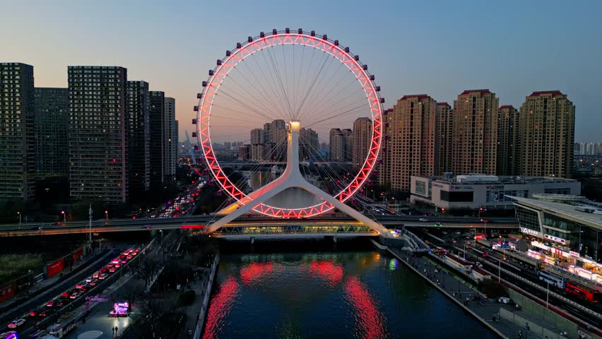 Aerial view of the illuminated Tianjin Eye reflecting in the river, contrasted against the city