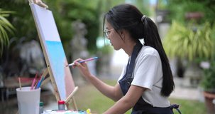 young asian woman adding detail to blue sky on canvas during relaxing outdoor painting session holding brush with gentle posture surrounded by green plants in quiet garden space - Powered by Shutterstock - Get 15% off with code: PIKWIZARD15