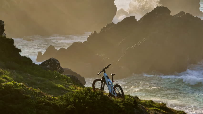 Mountain bike resting on a green, grassy coastal cliff overlooking the crashing waves and rocky formations of the Atlantic Ocean at sunset in Portugal, representing adventure and challenging trails