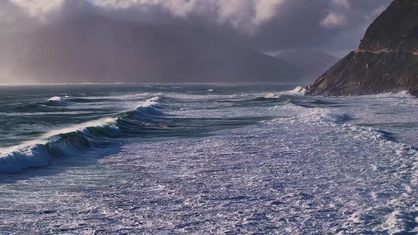 Aerial view of the coastline with waves crashing onto the shore under a cloudy sky, creating a dynamic interplay of light and shadow, Cape Town, Western Cape, South Africa.