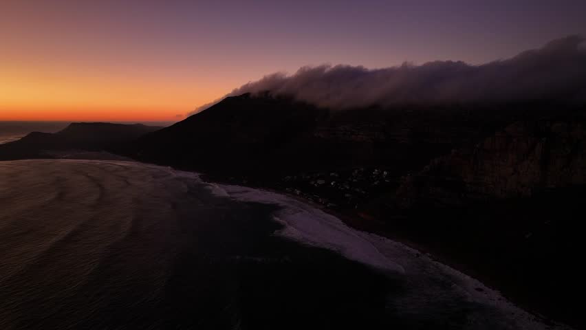 Aerial view of a dark coastline meeting the ocean, where the surf crashes onto the beach near rugged, shadowy mountains at dusk, Cape Town, Western Cape, South Africa.