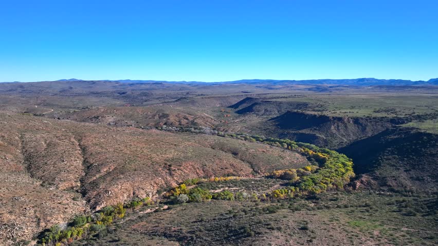 Aerial view of a winding river lined with vibrant trees cutting through the arid landscape under a clear blue sky, Agua Fria, Arizona, United States.