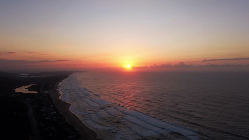 Aerial view of the sun setting over the ocean, casting a warm glow on the waves and coastline, Wilderness, Western Cape, South Africa.