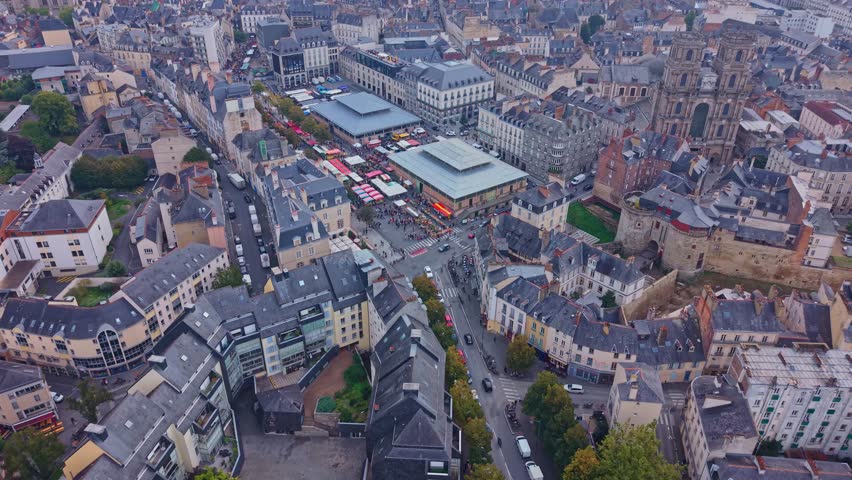 Place des Lices during busy market day with stalls crowds and historic buildings, aerial of birds soaring over car traffic in urban center. Rennes, Brittany