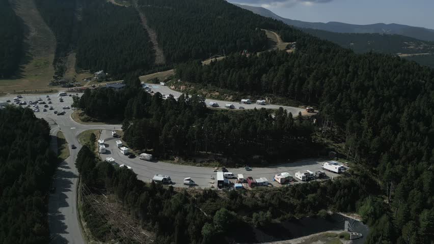 High-angle drone footage capturing a curved mountain parking area filled with motorhomes and campers, surrounded by dense evergreen forests and rolling hills under a clear sky