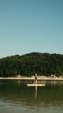 Young woman paddling on a SUP board across a calm lake surrounded by hills during a peaceful summer sunset. 4k vertical footage