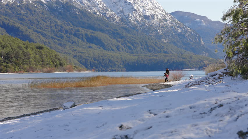 Mother and daughter skip stones into a cold river in Patagonia, Argentina, surrounded by snowy riverbanks, winter vegetation, calm flowing water, and forested Andean foothills, bonding concept