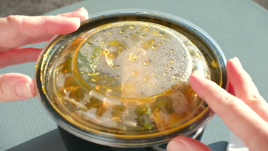 Hands open plastic lid from hot takeout container asian noodle soup ramen with chicken and vegetables, ready to eat for delicious, convenient lunch on sunny day. Hands open takeout container of ramen