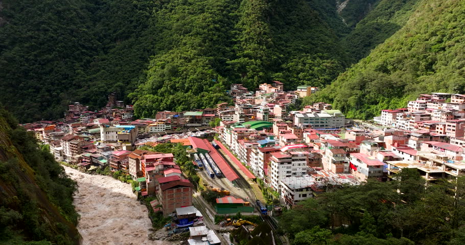 Aerial establishing orbit of Aguas Calientes town nestled between steep green mountains near Machu Picchu, roaring river