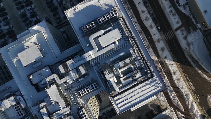 Top-down drone footage of a building rooftop in winter, showing snow-covered HVAC units, ventilation ducts, pipes, and industrial infrastructure in an urban setting.