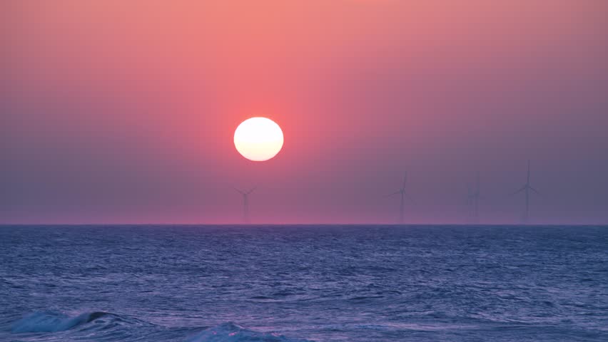 A vibrant sunset casts a warm glow over the Taiwan Strait, with the sun dipping towards the horizon. Offshore wind turbines stand silhouetted against the colorful sky in Zhunan.