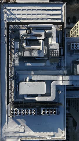 Top-down drone footage of a building rooftop in winter, showing snow-covered HVAC units, ventilation ducts, pipes, and industrial infrastructure in an urban setting.