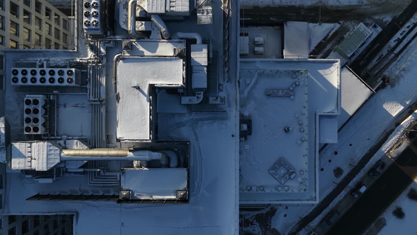 Top-down drone footage of a building rooftop in winter, showing snow-covered HVAC units, ventilation ducts, pipes, and industrial infrastructure in an urban setting.