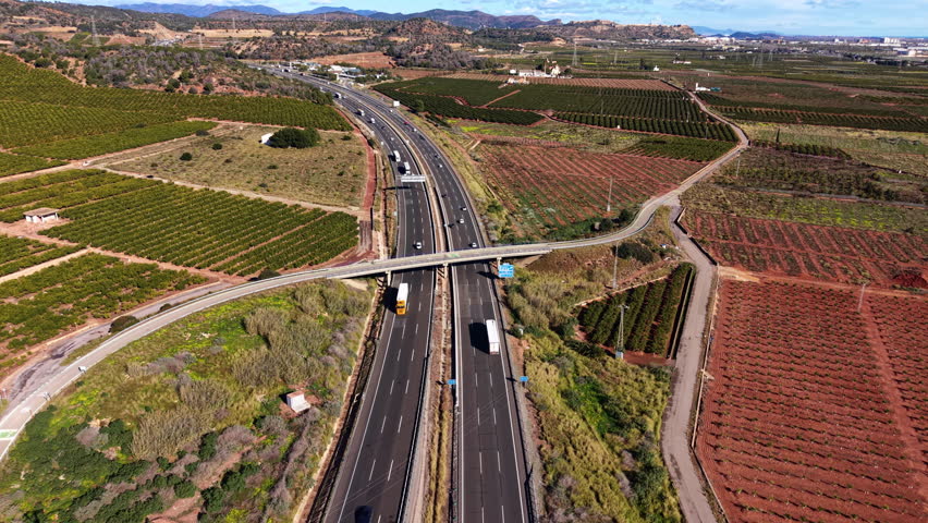 Aerial view of a multilane highway intersected by a rural overpass, surrounded by patterned agricultural fields and moving traffic in the Valencia countryside under clear skies