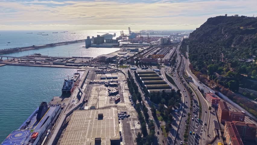 Industrial and commercial Port of Barcelona, showing a large ferry, shipping containers, cranes, and the Montjuïc hill in the background, Spain. Aerial drone forward