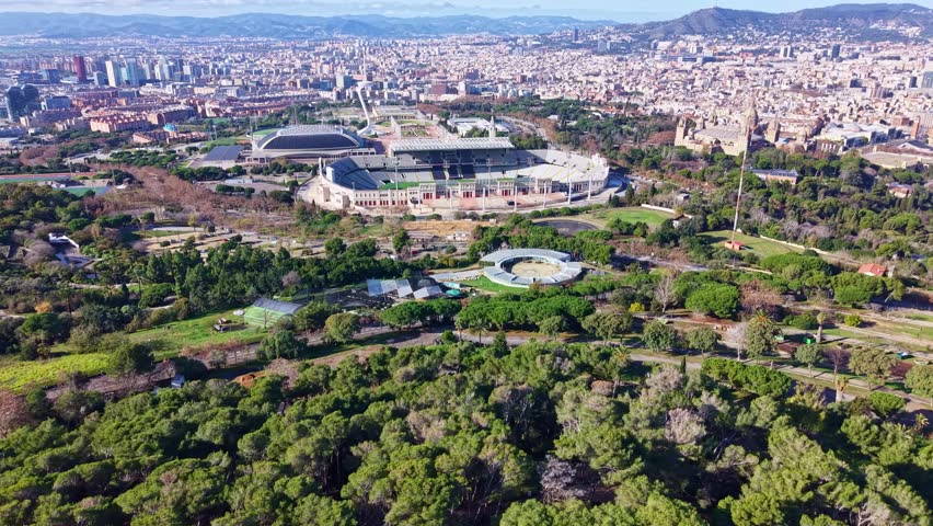 Lluís Companys Olympic Stadium nestled in the green landscape of Montjuïc hill, with the sprawling Barcelona cityscape in the background, Spain. Aerial forward