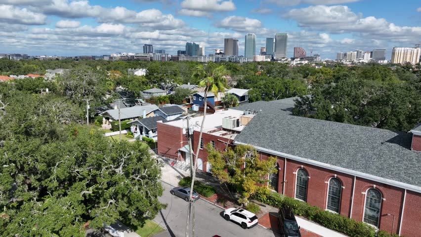 Hyde Park neighborhood in Tampa, Florida. Lush tree-lined streets, residential buildings and palm trees. Downtown Tampa skyline in distance. Aerial forward wide shot. Walking pedestrian on sidewalk.
