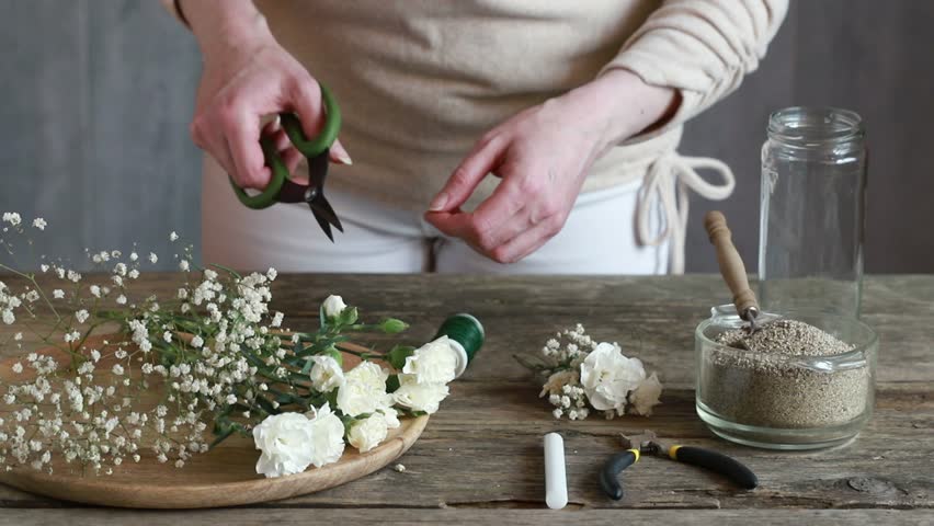Florist at work: woman shows how to make romantic candle holder using a glass jar, carnations flowers, gypsophila paniculata twigs and sand. Step by step, tutorial. Images available.