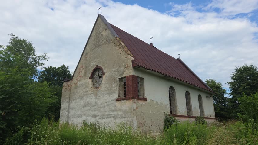Abandoned plastered church ruin with a red roof and round window, surrounded by wild green grass and trees under a cloudy blue sky on a sunny summer day