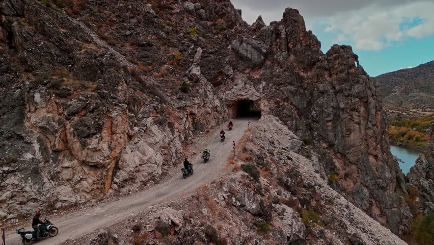 Aerial view of a motorcycle convoy entering a tunnel on a narrow cliff road in Karanlik Canyon beside the Euphrates River in Turkey. Dramatic rocky canyon landscape for travel and adventure concepts.