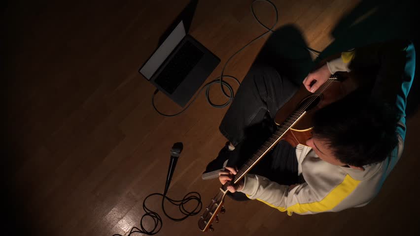 Musician sitting on a wooden floor, playing an acoustic guitar while a laptop and microphone are set up for home recording, focusing on music production and creativity