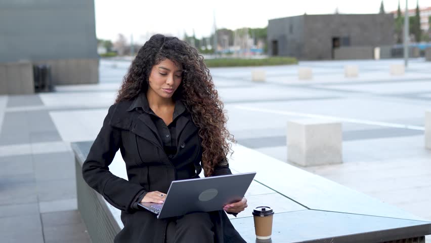 Curly-haired young woman, who is a professional, sits on a bench in a contemporary city square as she works on her laptop. She smiles warmly and greets someone with a wave