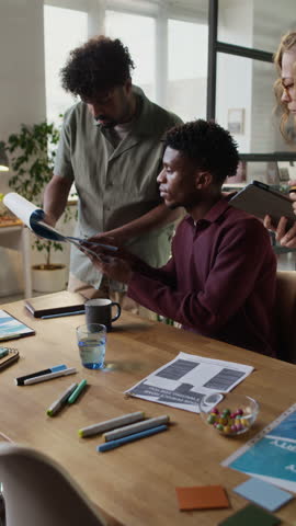Vertical shot of young African American man helping his colleagues with tasks while sitting at table in modern office