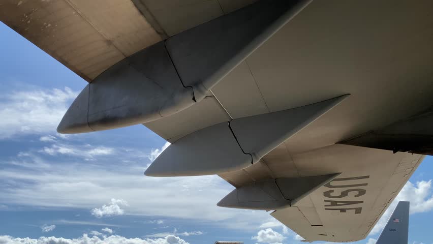 A jet airplane wing stretches across a bright blue sky dotted with clouds. Aerodynamic fairings and panels are seen along the underside of the aircraft wing in daylight.
