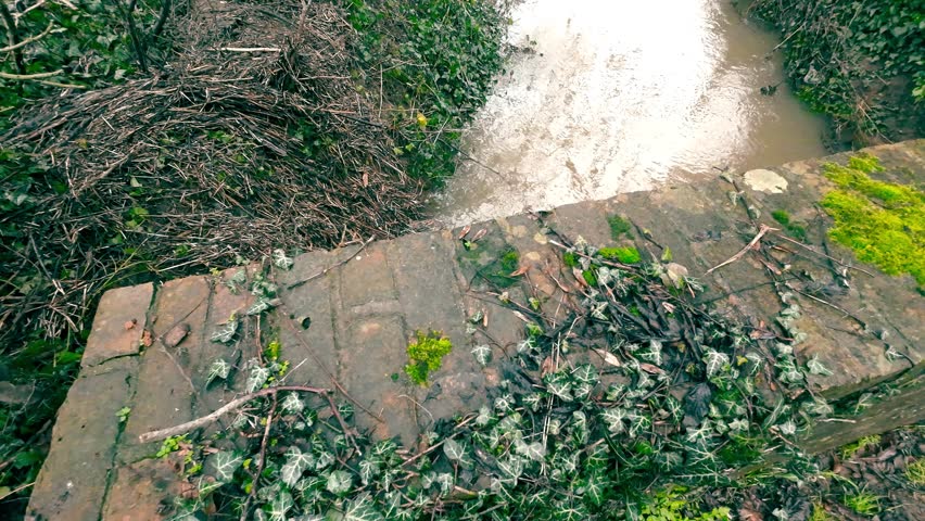 High angle view of a narrow stream flowing under an old mossy brick bridge next to a muddy country road