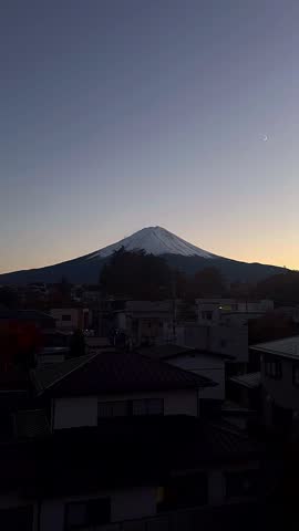 snow capped mt fuji rises above quiet residential rooftops at dusk, framed by a soft twilight sky and a delicate crescent moon, creating a peaceful evening scene in japan.